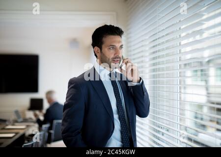 Uomo d'affari con bearded che indossa il vestito e la cravatta in piedi vicino ad una finestra, usando il telefono mobile. Foto Stock
