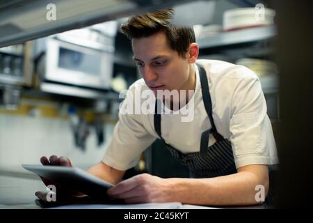Chef con grembiule blu in cucina, utilizzando un tablet digitale. Foto Stock