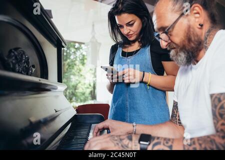 Uomo tatuato bearded con capelli lunghi e bicchieri che suonano piano, donna che tiene il telefono cellulare ascolto. Foto Stock