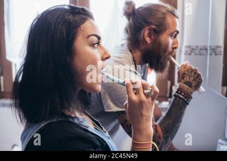 Uomo tatuato con lunghi capelli e donna con lunghi capelli castani in piedi davanti allo specchio, spazzolando i denti. Foto Stock