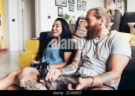 Uomo tatuato con lunghi capelli bruna e donna con lunghi capelli marroni seduti su un divano, sorridente mentre si gioca a console. Foto Stock