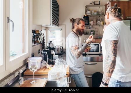 Due uomini tatuati con bearded con lunghi capelli di brunetta in piedi in una cucina, mangiare. Foto Stock