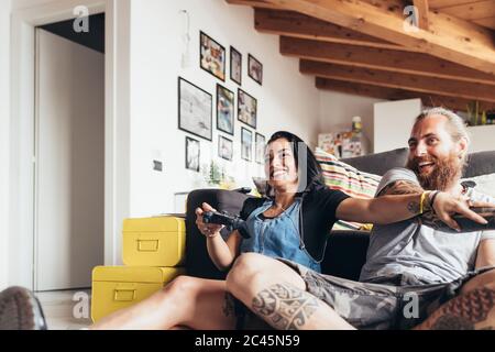 Uomo tatuato con lunghi capelli bruna e donna con lunghi capelli marroni seduti su un divano, sorridente mentre si gioca a console. Foto Stock