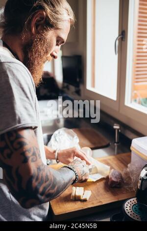 Uomo tatuato con lunghi capelli di brunetta in piedi in cucina, preparando il cibo. Foto Stock