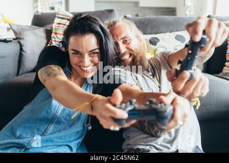 Uomo tatuato con lunghi capelli bruna e donna con lunghi capelli marroni seduti su un divano, sorridente mentre si gioca a console. Foto Stock