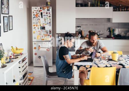 Uomo tatuato con lunghi capelli brunette e donna con lunghi capelli marroni seduti a un tavolo da cucina, utilizzando un computer portatile e un telefono cellulare. Foto Stock