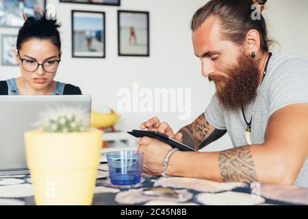 Uomo tatuato con lunghi capelli brunette e donna con lunghi capelli marroni seduti a un tavolo da cucina, utilizzando un computer portatile e un telefono cellulare. Foto Stock