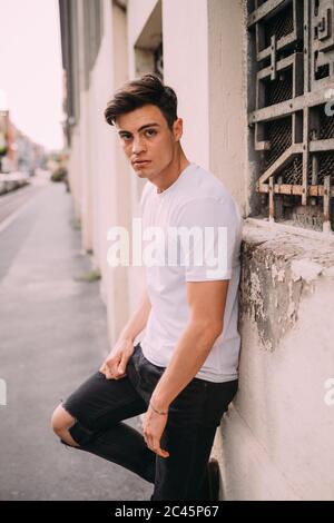 Ritratto di giovane uomo con capelli castani scuri con T shirt bianca e jeans neri strappati appoggiati alla parete Foto stock Alamy