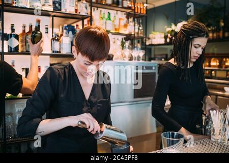 Due giovani donne che indossano abiti neri in piedi dietro il bancone bar, preparando le bevande. Foto Stock