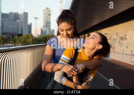 Due giovani donne con lunghi capelli marroni in piedi sul ponte urbano, abbraccianti e sorridenti. Foto Stock