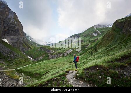 Uomo turistico con zaino in piedi su un sentiero roccioso di montagna nella verde valle collinare. Alpinista che gode della vista di splendide alte rocce e scogliere mentre si viaggia da solo. Concetto di escursioni, viaggi Foto Stock