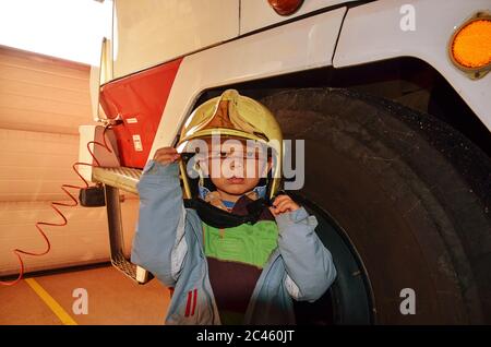 Ragazzino che agisce come un pompiere. Ragazzo in piedi accanto a una vera macchina vigile del fuoco. Felice adorabile bambino con cappello pompiere accanto camion fuoco rosso Foto Stock