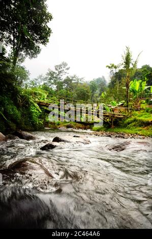 Paesaggio tropicale con fiume roccioso sotto il ponte di legno e tipica casa rurale indonesiana nella giungla di Giava occidentale Foto Stock