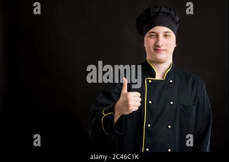 Ritratto di giovane maschio vestito in un vestito nero da chef che mostra pollici in posa su uno sfondo nero isolato con area pubblicitaria spazio di copia Foto Stock