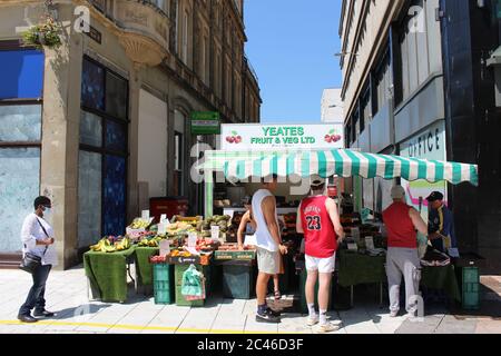 Una fotografia di un commerciante di frutta e verdura su Cardiff Queen Street. I clienti che seguono misure di social distancing in coda. Mercato agricolo. Foto Stock