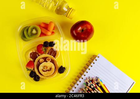 Cibo gustoso in contenitore e matite colorate su sfondo giallo. Concetto di ritorno a scuola. Foto Stock