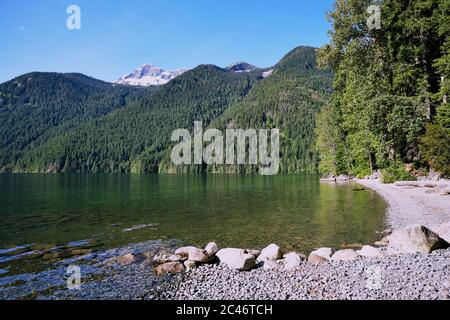 Spiaggia e varo di barche al lago Chilliwack (Sxotsaquel), BC. Torreggianti su pendii boschivi con conifere sono alte montagne con pareti rocciose esposte e. Foto Stock