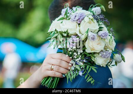 Mano della sposa che tiene bouquet di fiori sulla spalla dello sposo Foto Stock