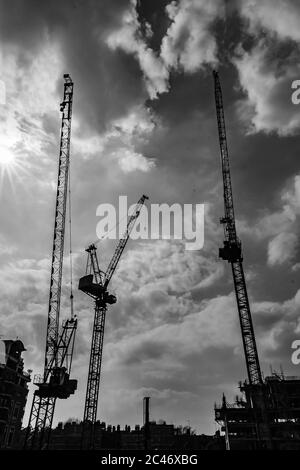 Immagine in scala di grigi ad angolo basso di gru da costruzione che lavorano sotto a. cielo nuvoloso a Londra Foto Stock