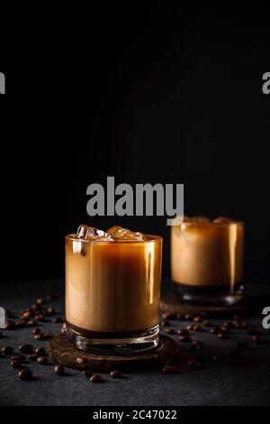 Una foto verticale di 2 bicchieri di roccia con caffè freddo ghiacciato con latte, chicchi di caffè intorno, sfondo scuro, ombre profonde Foto Stock