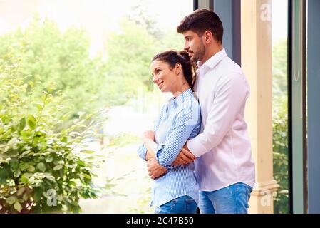 Un colpo di coppia felice in piedi alla finestra e godendosi la compagnia dell'altro. Bell'uomo giovane che abbracciava la sua ragazza mentre si rilassava a casa. Foto Stock