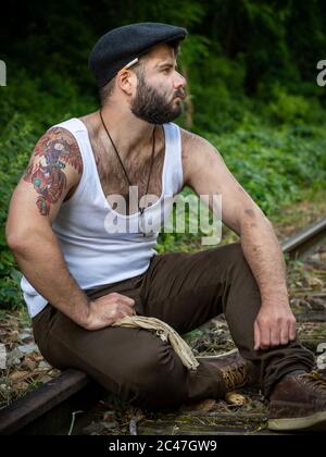 Giovane uomo francese con tatuaggi e macchie di carbone sulla pelle, portato fuori su una pista ferroviaria in disuso con luce naturale a Parigi, Francia Foto Stock