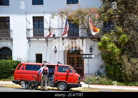 Vista posteriore di un uomo anziano che carica una bicicletta nella Volkswagen Vanagon rossa d'epoca, di fronte a un bellissimo edificio in stile spagnolo a Santa Barbara, CA, USA Foto Stock