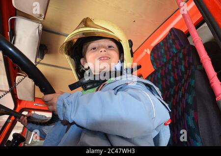 Ragazzino che agisce come un pompiere. Ragazzo seduto in una vera macchina vigile del fuoco. Felice adorabile bambino ragazzo con cappello vigile del fuoco seduto in camion fuoco rosso guardando fuori Foto Stock