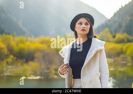 Bella donna in un cappotto bianco e cappello nero che posa su uno sfondo di montagne autunnali Foto Stock