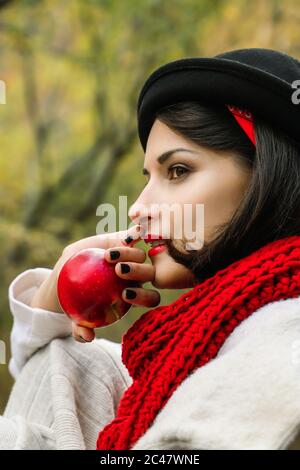 Bella donna con cappello nero e sciarpa rossa con una mela rossa in mano. Picnic all'aperto in autunno Foto Stock