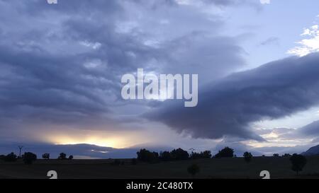 Vista di un cielo nuvoloso scuro su un campo in pendenza con pochi alberi Foto Stock