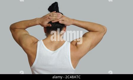 Giovane ispanico con i capelli raccolti fiocco in t-shirt bianca senza maniche, visto dal retro, con le mani insieme sulla nuca sullo sfondo grigio chiaro Foto Stock