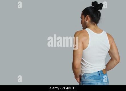 Giovane maschio ispanico con i capelli raccolti fatto arco in t-shirt bianca senza maniche, visto da dietro su sfondo grigio chiaro Foto Stock