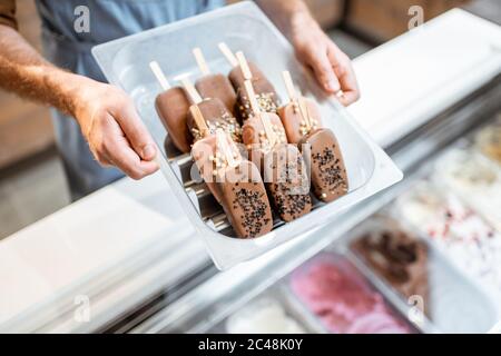 Venditore che tiene un vassoio con gelato al cioccolato su un bastone nel negozio, primo piano su gelato su uno sfondo blu grembiule Foto Stock