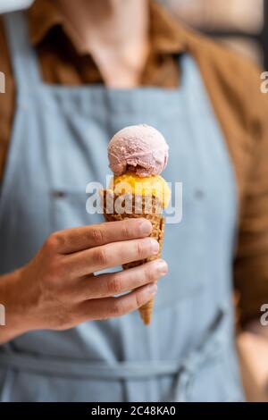 Venditore in grembiule che tiene il gelato in un cono del waffle, primo piano. Vendita di gelato in un negozio Foto Stock