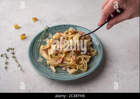 La mano della donna tiene una forchetta con fettuccine di pasta con funghi, pancetta e parmigiano in piatto su uno sfondo di legno chiaro, primo piano Foto Stock