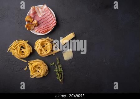 Vista dall'alto ingredienti di pasta fettuccine con funghi, pancetta e parmigiano. Cibo grezzo su sfondo di grafite scuro con spazio di copia Foto Stock