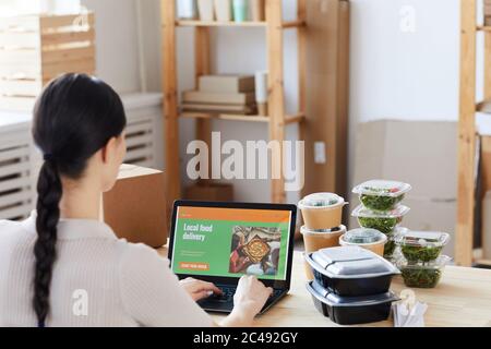 Vista posteriore della giovane donna seduta al tavolo con scatole di cibo e ordinare online cibo in ufficio Foto Stock