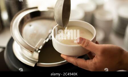 Una mano di mans tiene un mestolo e mette sulla zuppa, UN uomo che versa la zuppa in piatto bianco dal tureen Foto Stock