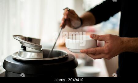 Una mano di mans tiene un mestolo e mette sulla zuppa, UN uomo che versa la zuppa in piatto bianco dal tureen Foto Stock