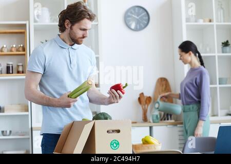 Giovane uomo che disimballa verdure fresche dalla scatola con donna in piedi sullo sfondo sono in cucina Foto Stock