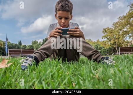 Concentrato marrone capelli ragazzo seduto al parco giochi con uno smartphone. Foto Stock