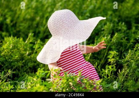 Carino bambina in un vestito rosso e cappello cammina in estate a piedi nudi sull'erba verde, spazio per il testo Foto Stock