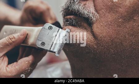 Primo piano di rasoio elettrico che è nelle mani di un barbiere.Serious master barba da taglio del cliente anziano nel salone. Foto Stock