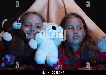 Bambini che indossano jammy rossi nel letto su sfondo nero. Ragazze con facce felici che giocano con i teddies. Amici che si divertono con i giocattoli in tenda coperta. Festa PJS per bambini. Concetto di tempo per bambini e divertimento. Foto Stock