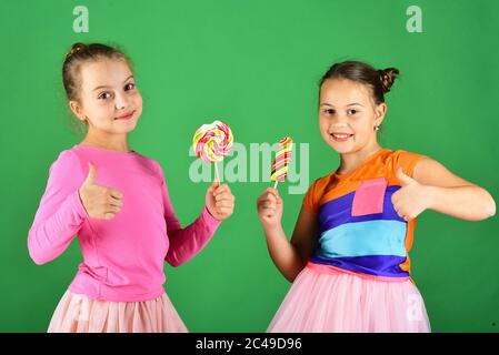 Le ragazze mangiano i caramelli dolci e colorati grandi. Concetto di pasticceria e infanzia. Le sorelle con lollipop rotondi e lunghi mostrano i pollici. I bambini con volti felici posano con caramelle su sfondo verde. Foto Stock