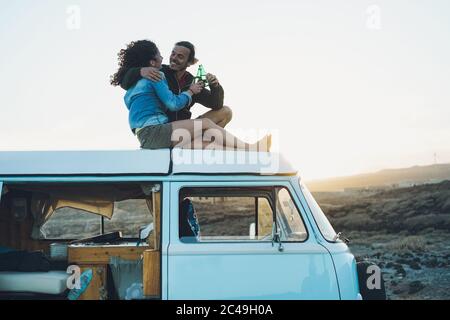 Coppia felice che si acclamano con le birre sul tetto minivan - Coppia di viaggio che si diverte sulla vacanza viaggio su strada - Amore, stile di vita van e concetto di vacanza Foto Stock