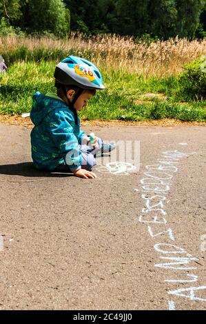 POZNAN, POLONIA - 30 maggio 2020: Ragazzo di due anni con elmetto di sicurezza con scrittura con pastello su un sentiero asfaltato nel parco Jan Pawla Foto Stock