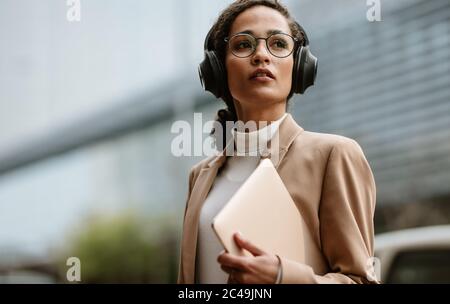 Uomo d'affari che indossa le cuffie che cammina lungo la strada della città tenendo un computer portatile. Donna d'affari che indossa cuffie e computer portatile in mano sulla strada della città. Foto Stock