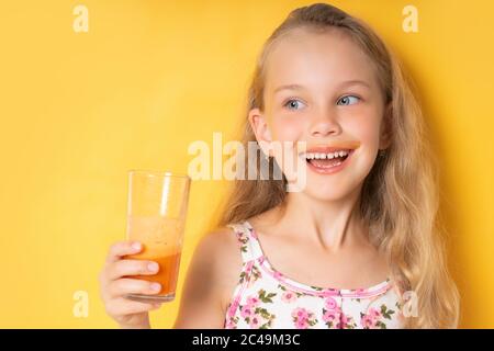 Sorriante bambina in abbigliamento estivo guardando la macchina fotografica che tiene in mano un bicchiere di succo di carota fresco. Girata in vita isolata in giallo, spazio per la copia Foto Stock